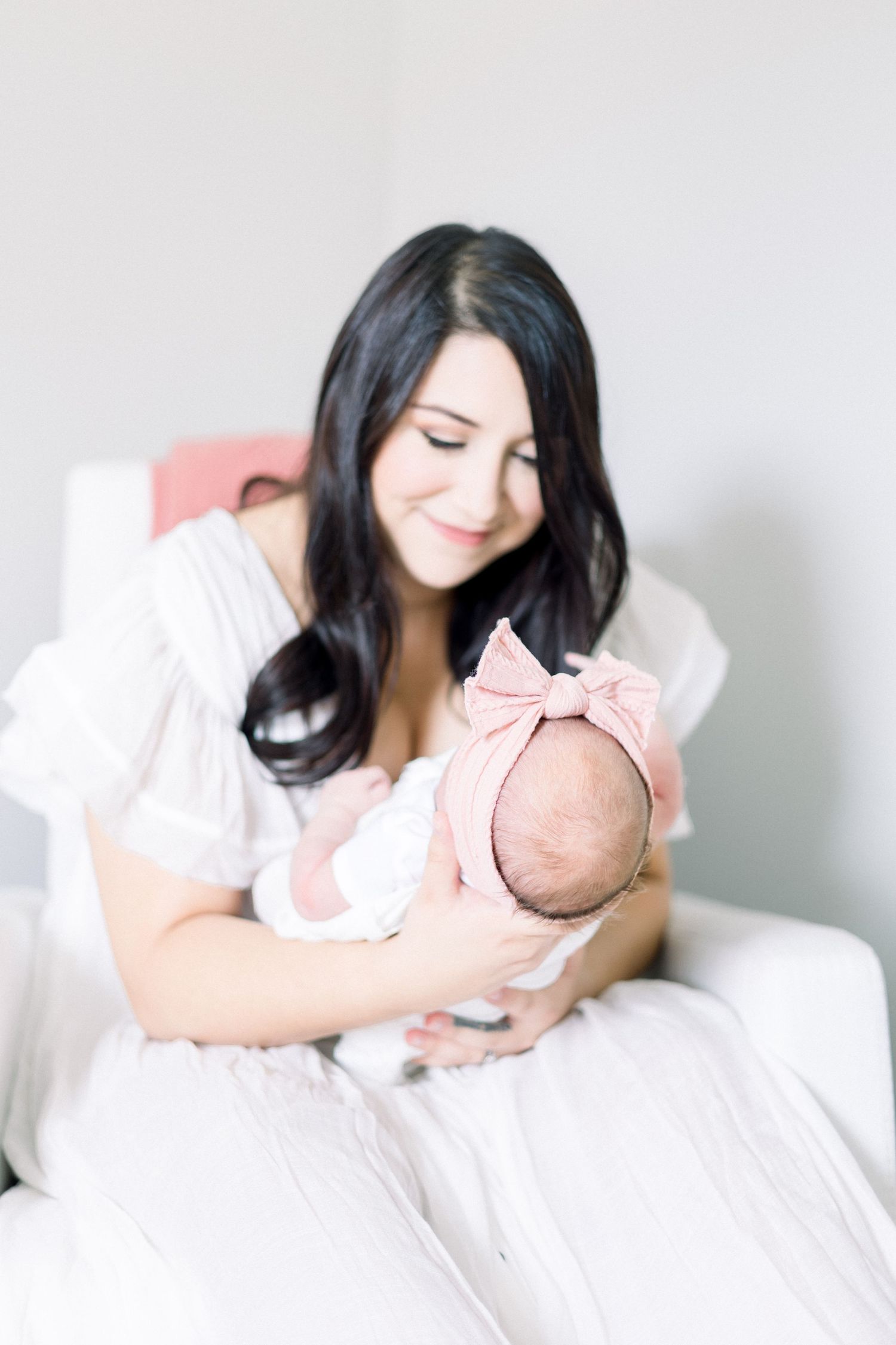 A person in white holds a sleeping newborn baby wearing a pink bow while sitting in a comfortable chair. Evans Newborn Photographer