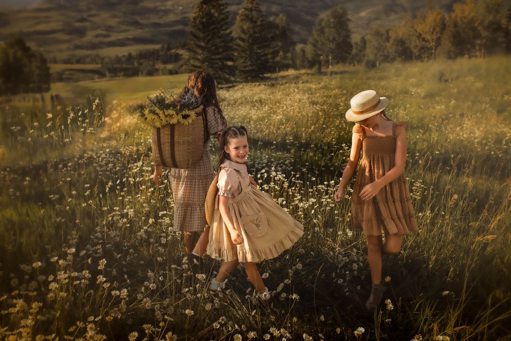 Three figures in brown dresses walking through a meadow of white flowers at golden hour.