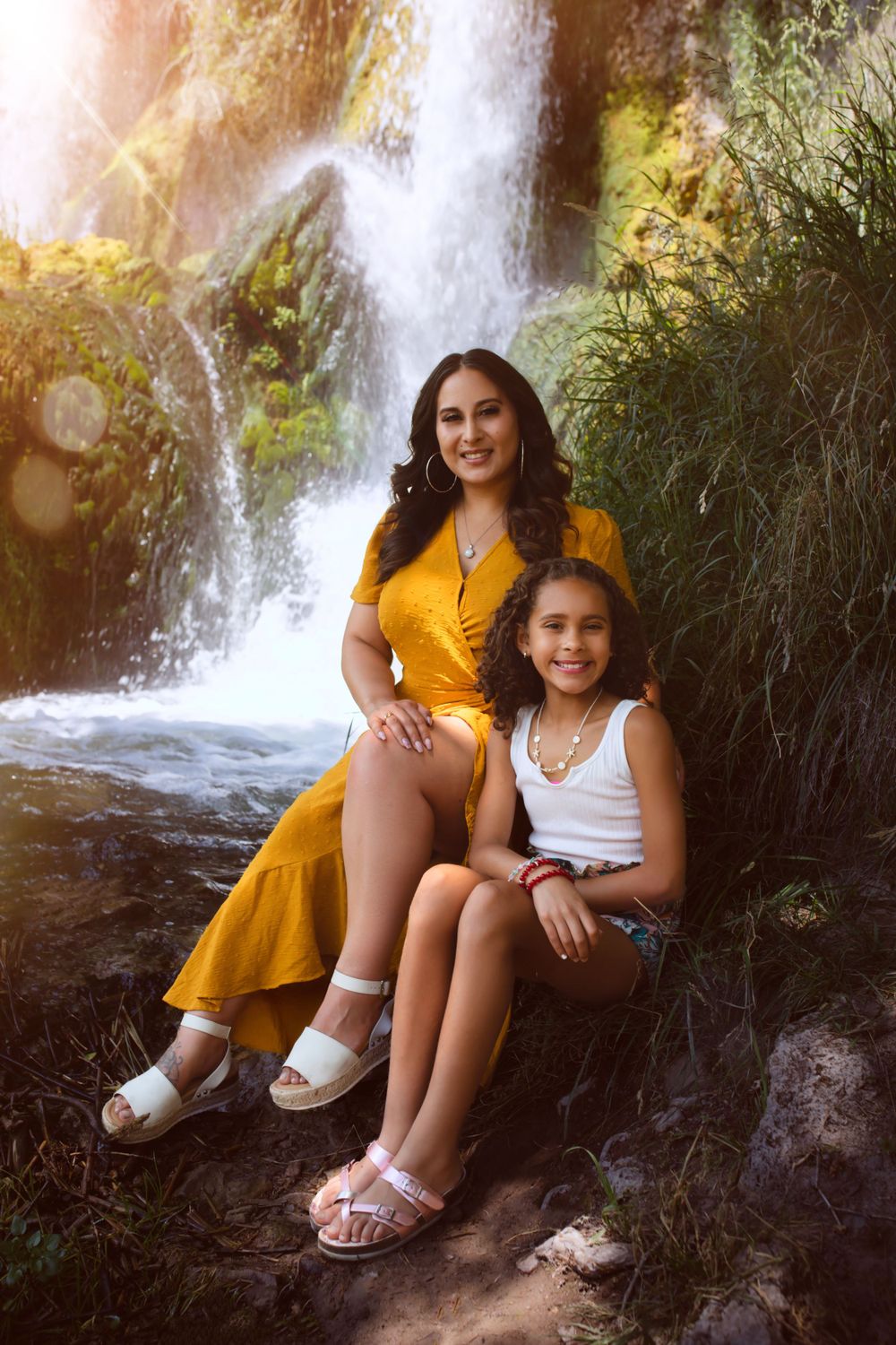 Mother and daughter sitting together by scenic waterfall on sunny day.
