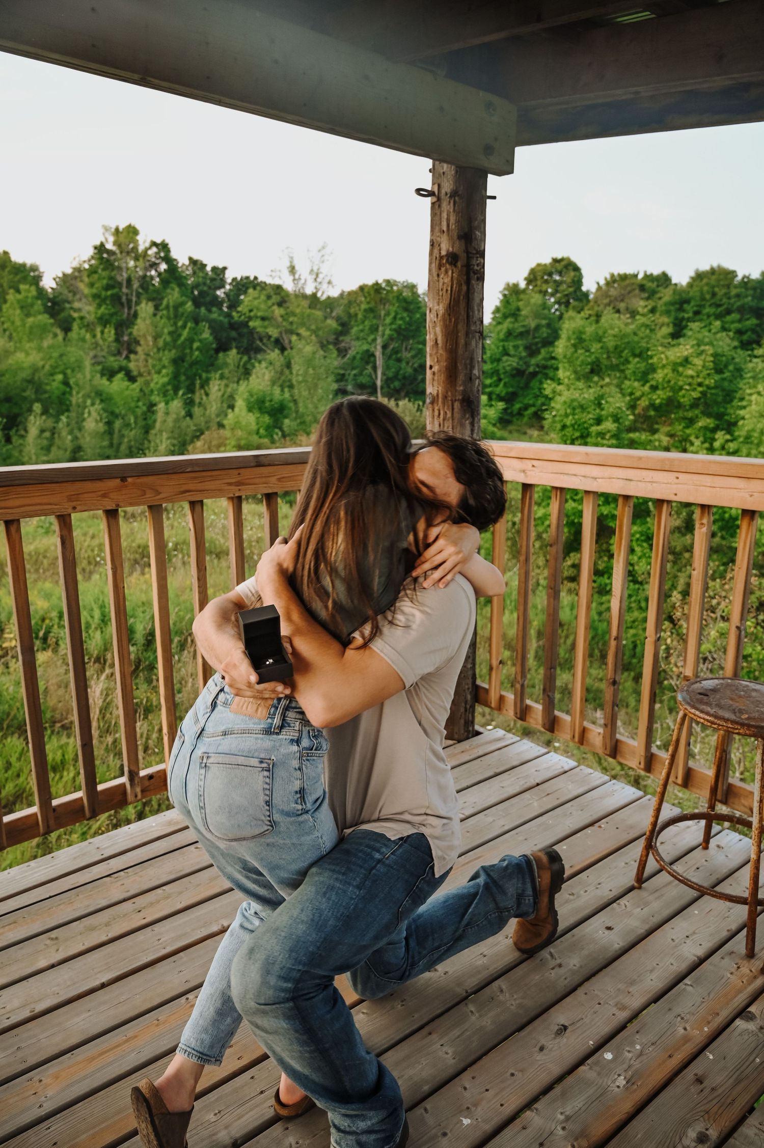 A couple embraces in a romantic moment on a wooden deck overlooking lush green trees and forest.