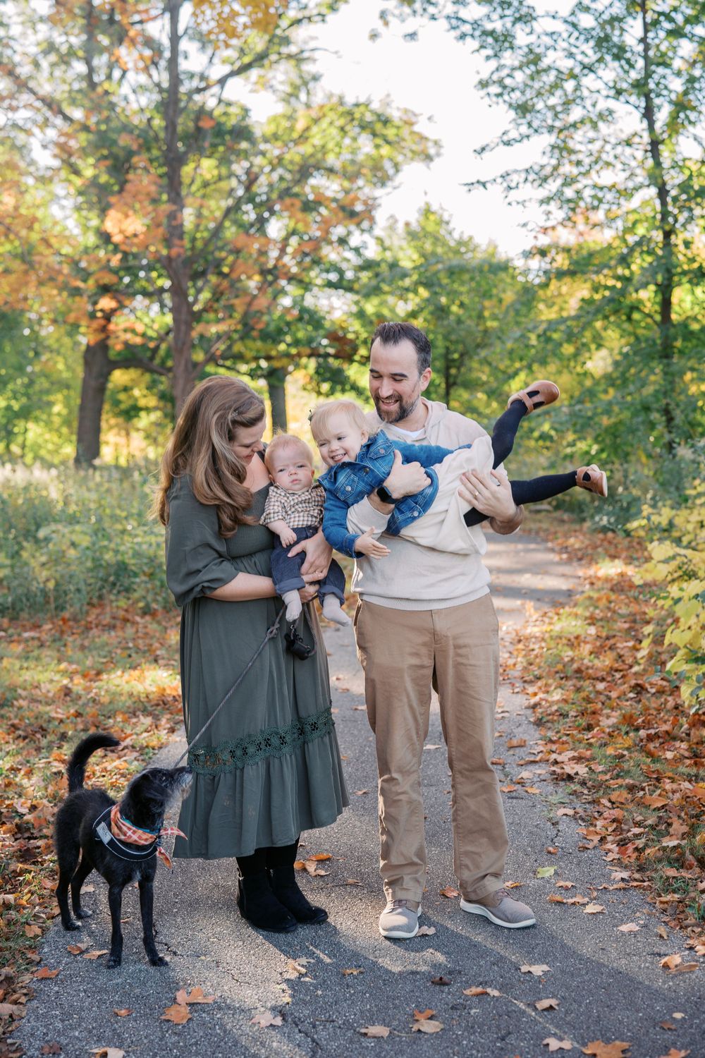 A family and their black dog enjoy an autumn stroll on a leaf-covered path surrounded by colorful trees.