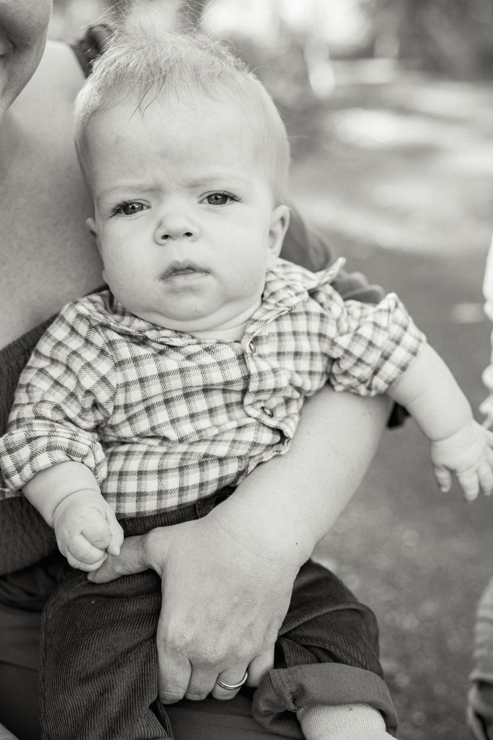 A baby wearing a checkered shirt sits outdoors in black and white photography.
