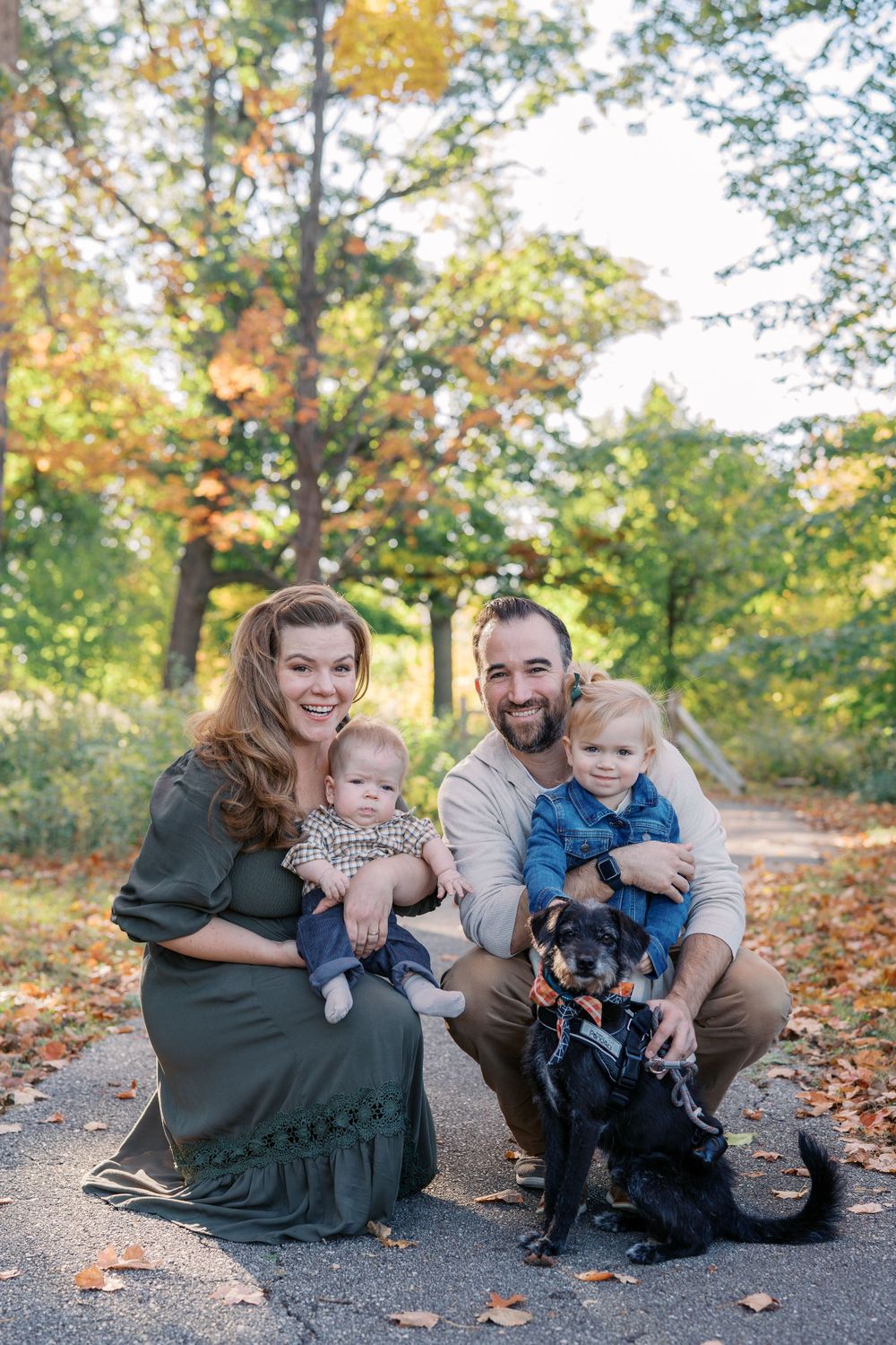 A family poses with their black dog on a fall path surrounded by autumn foliage.
