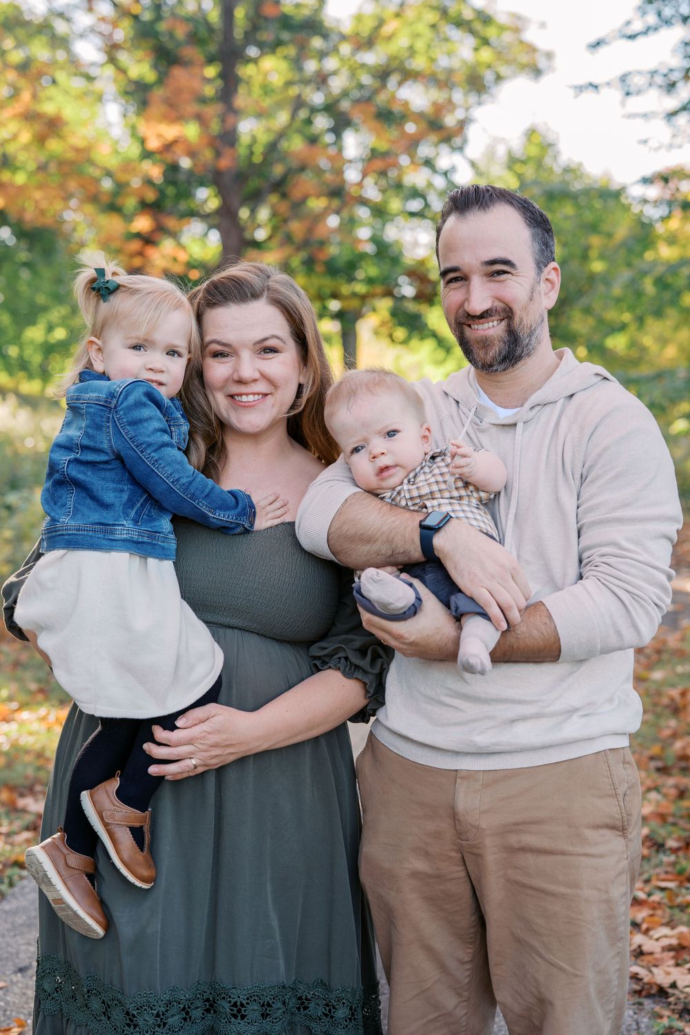 A family of four poses together outdoors on an autumn day with trees showing fall colors.