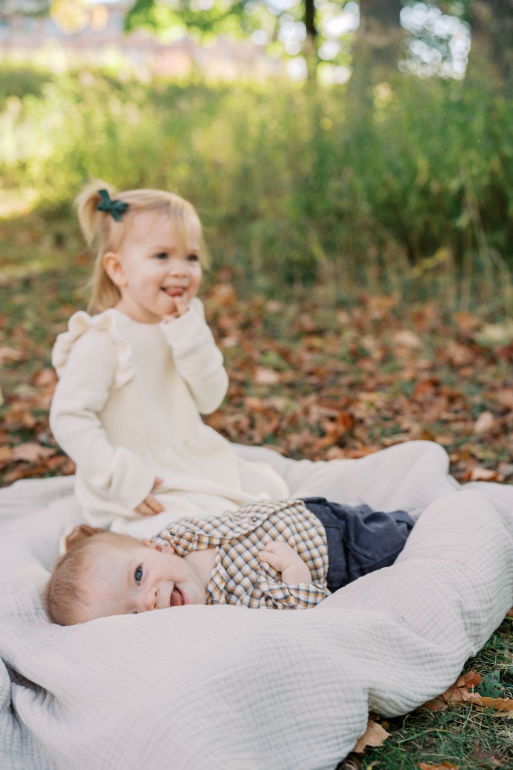 Two children share joyful moments on a white blanket surrounded by fallen autumn leaves.
