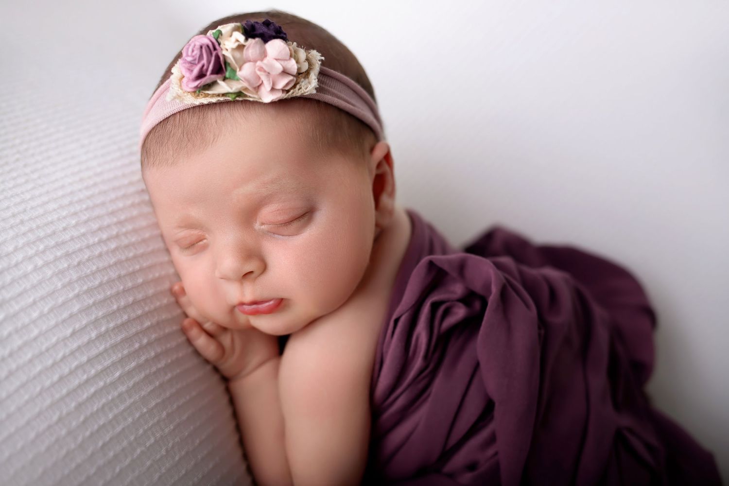 Sleeping newborn wrapped in soft purple fabric wearing delicate pink floral headband rests on white textured surface.