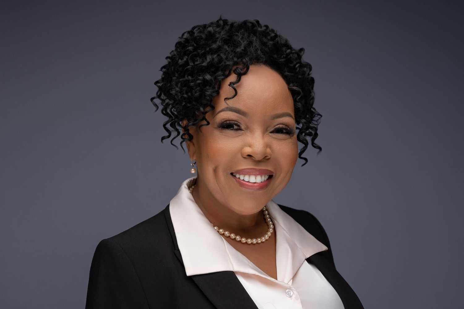 Professional business headshot of a person wearing a black blazer, white collared shirt and pearl necklace against grey background.