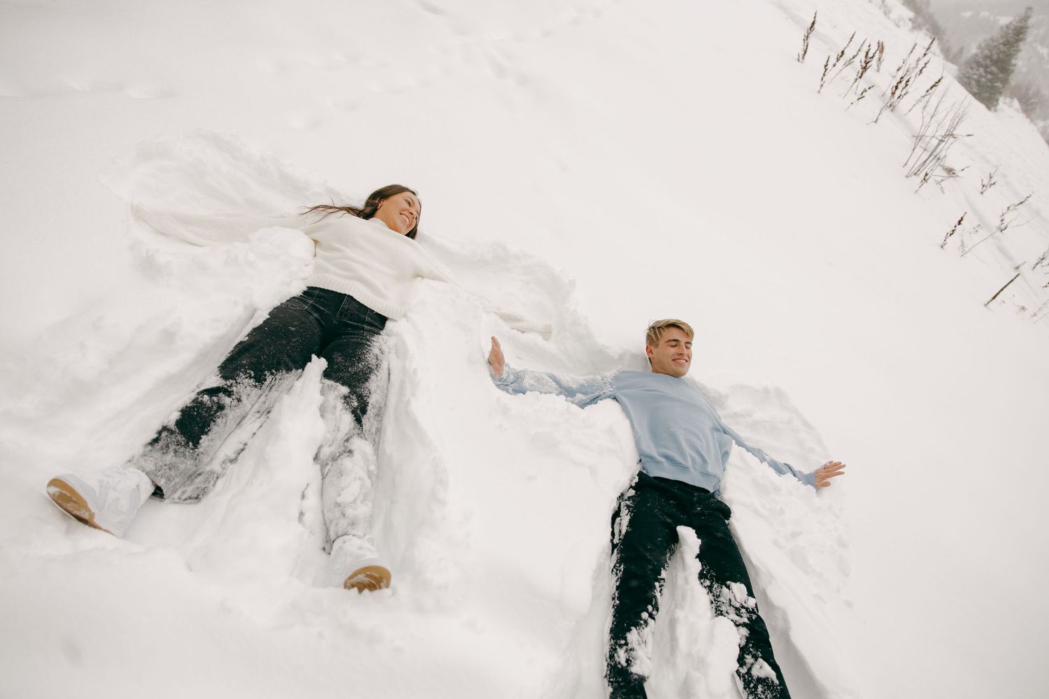 Couple laughing while making snow angels together on a snowy winter hillside.