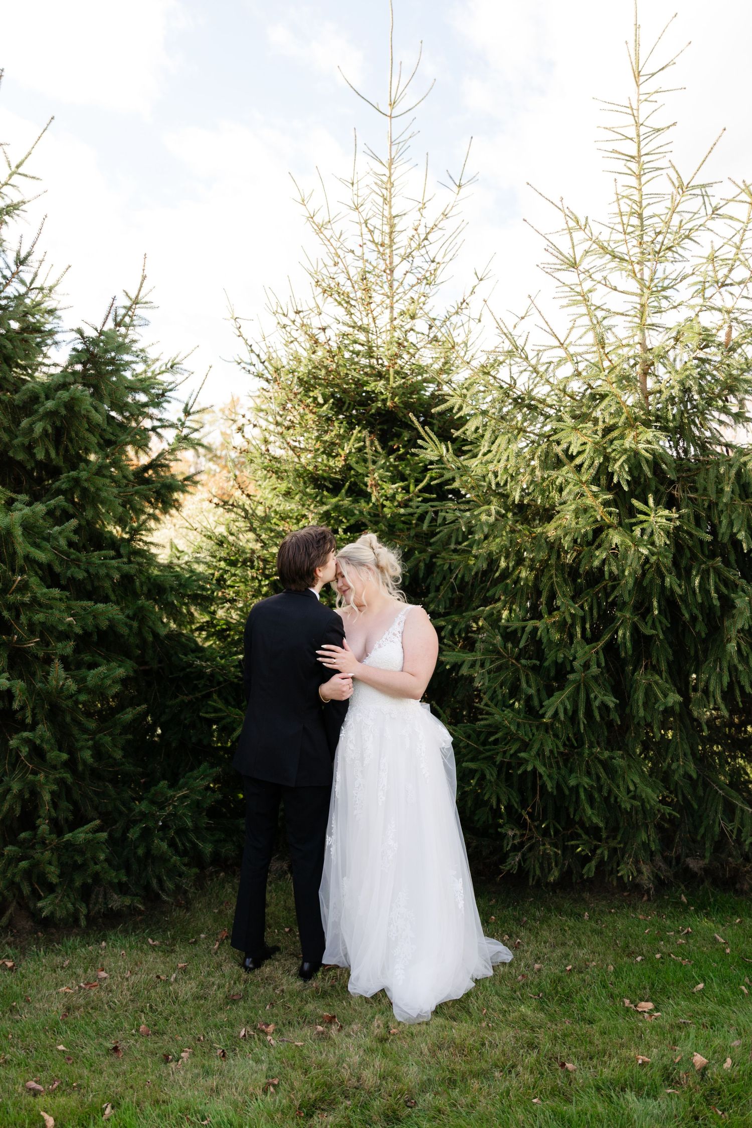 A romantic wedding couple shares an intimate moment in front of tall evergreen trees.