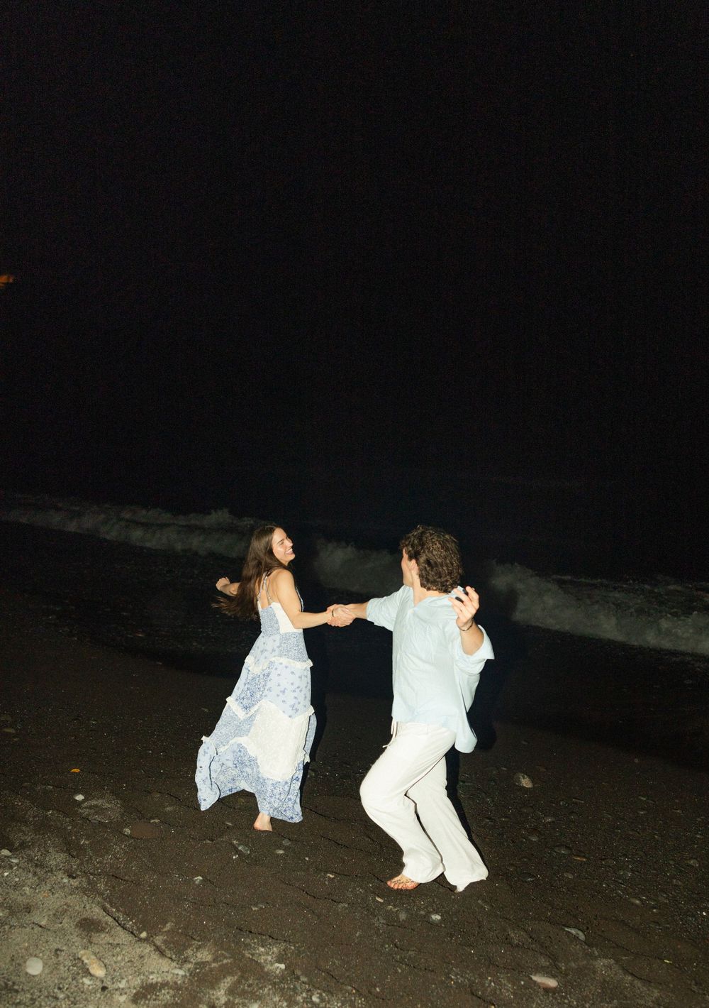 A couple in white outfits dances joyfully together on a dark beach at night.