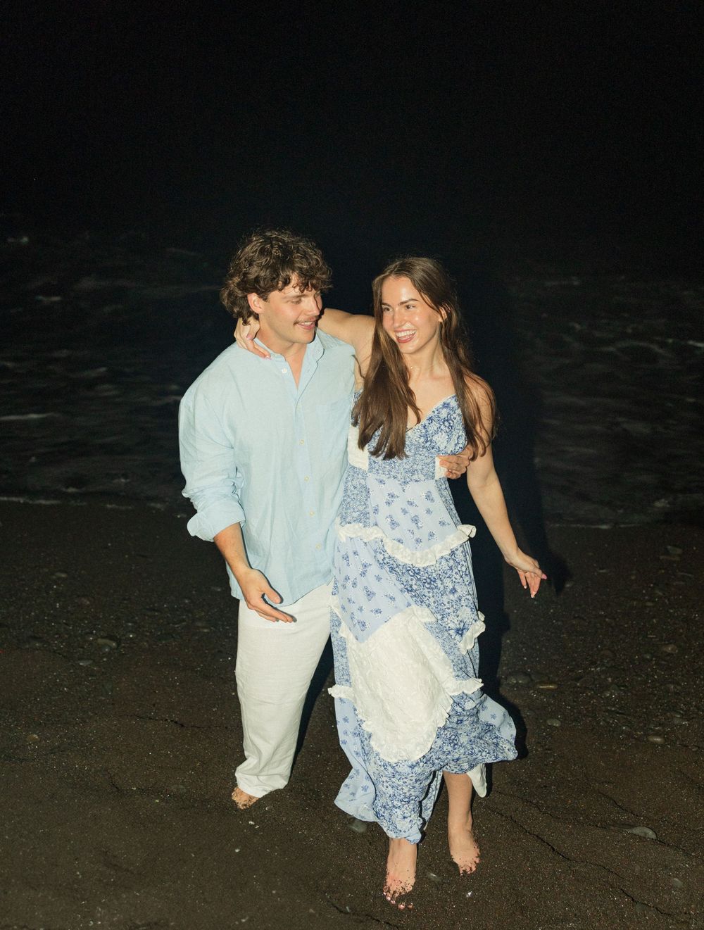 A couple in casual beach attire walks together at night along a dark sandy beach.