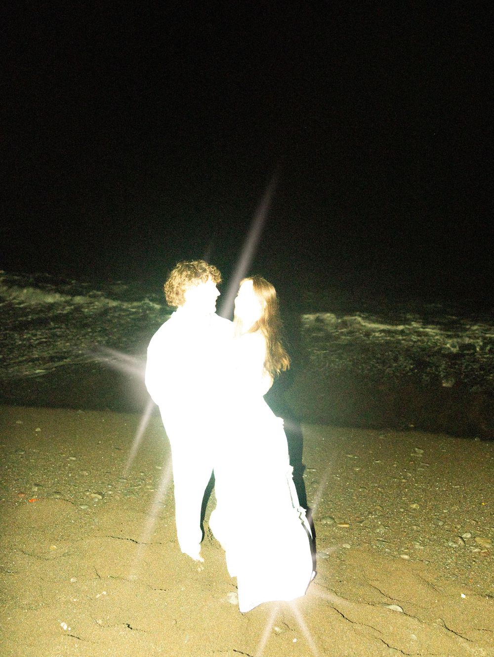 Two people in white clothing dance together on the beach with a bright camera flash illuminating them.