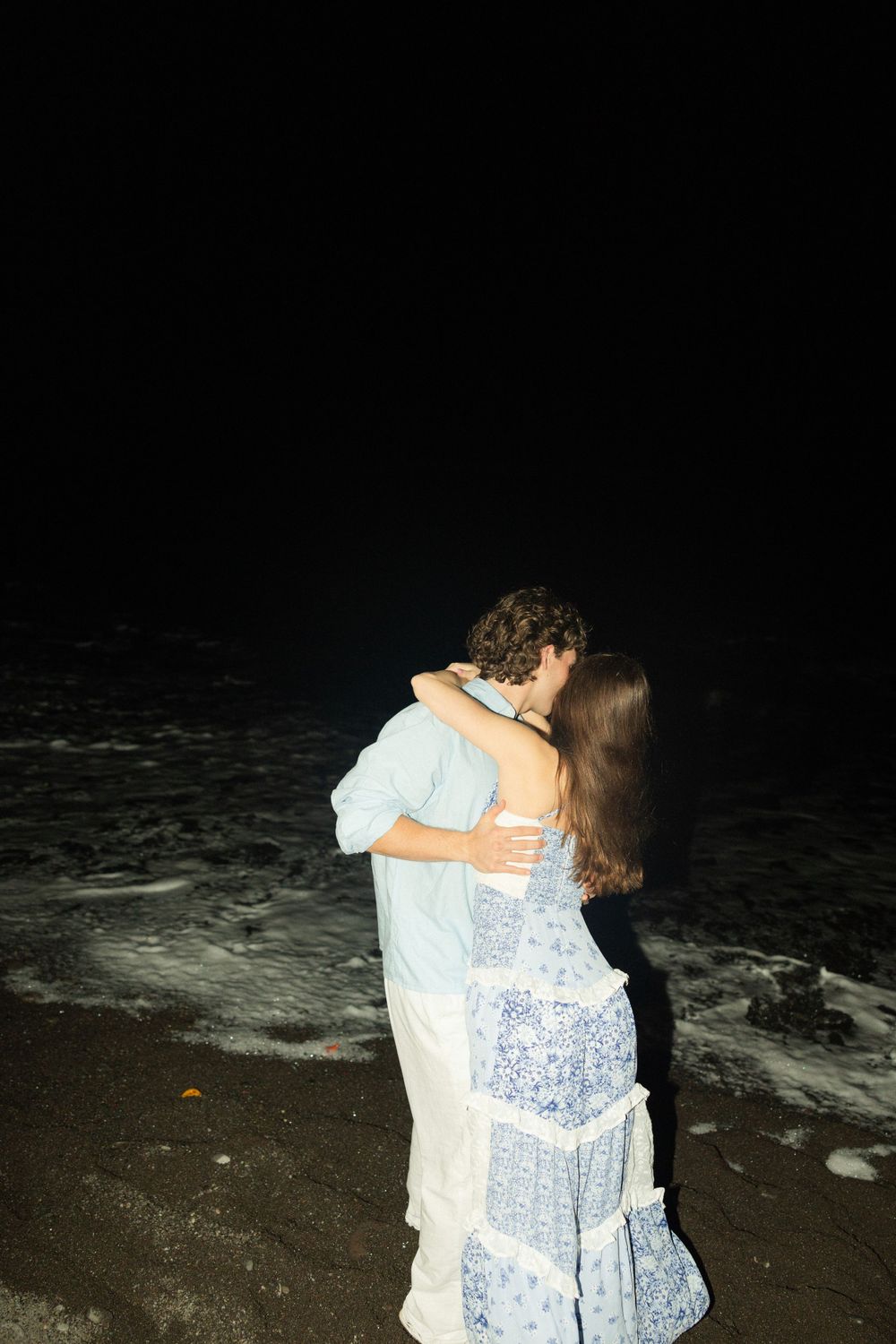 A romantic embrace captured at night on the dark shoreline.
