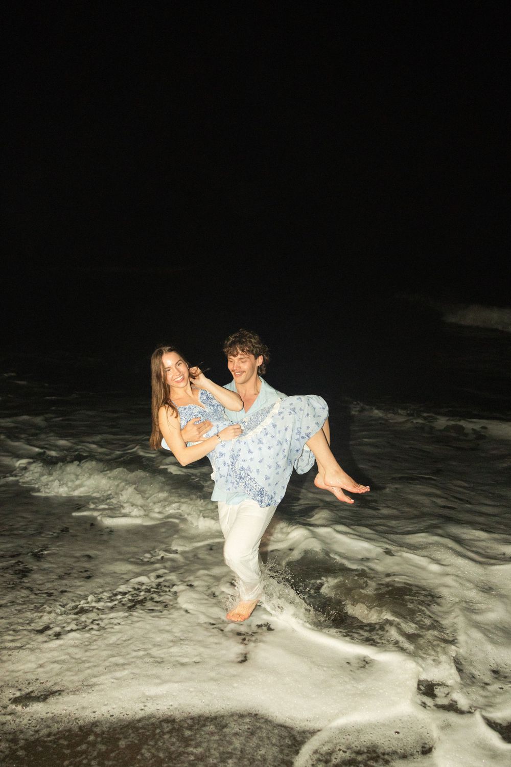 A couple plays in the ocean waves at night, with one person carrying the other in a playful embrace.