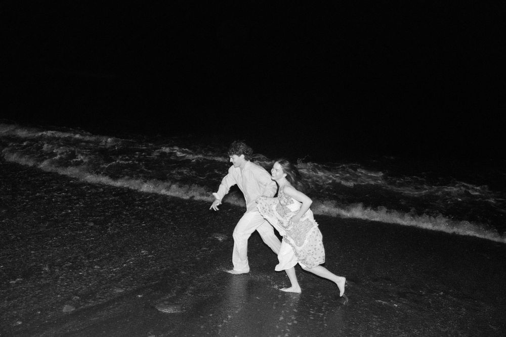 Two figures dressed in white run together along a dark beach at night next to crashing waves.
