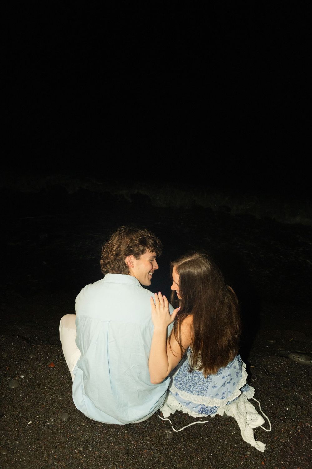 A couple sits close together on the beach sand at night.