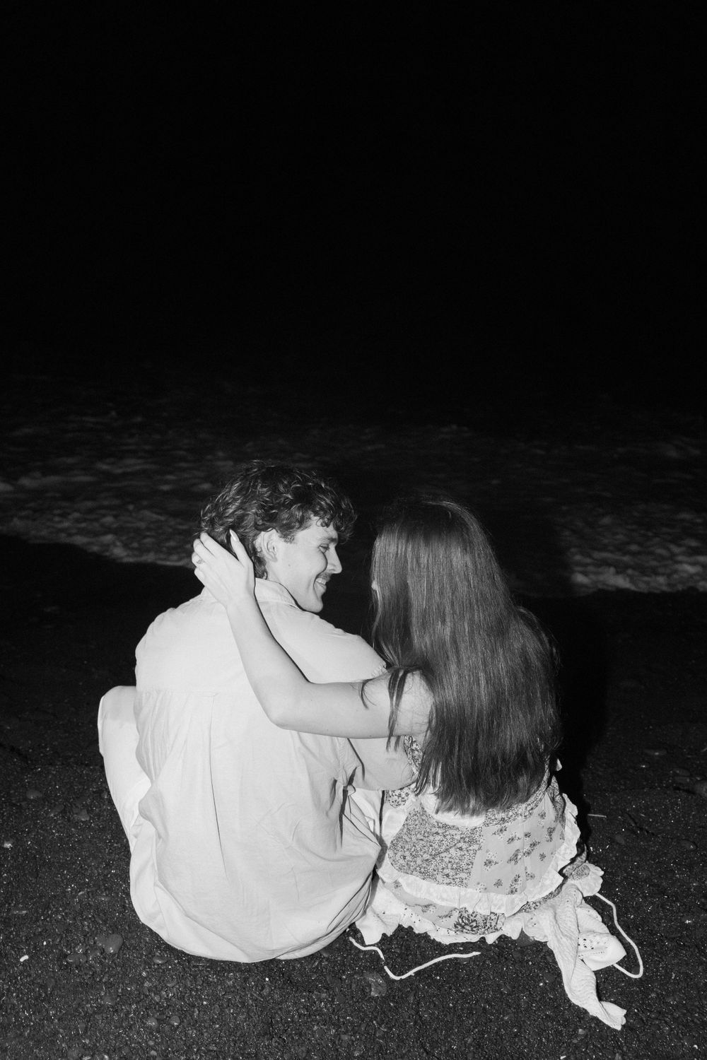 A couple sits together on a beach at night in black and white, facing the dark ocean waves.
