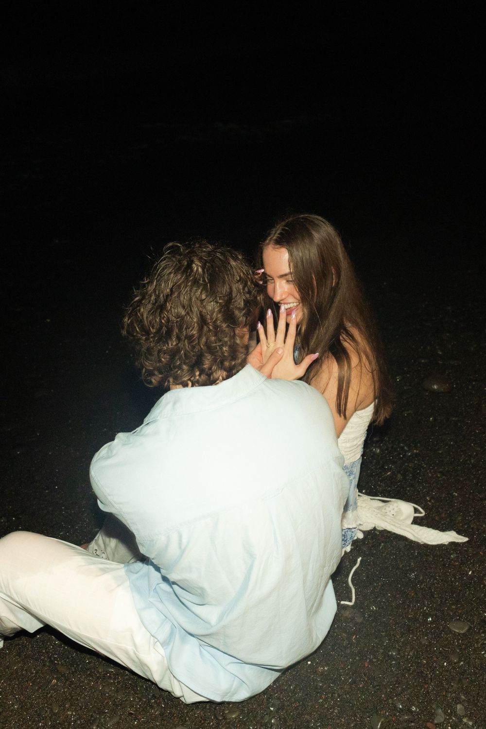 A couple sits together in dark lighting outdoors, one wearing white clothing while sharing an intimate moment.