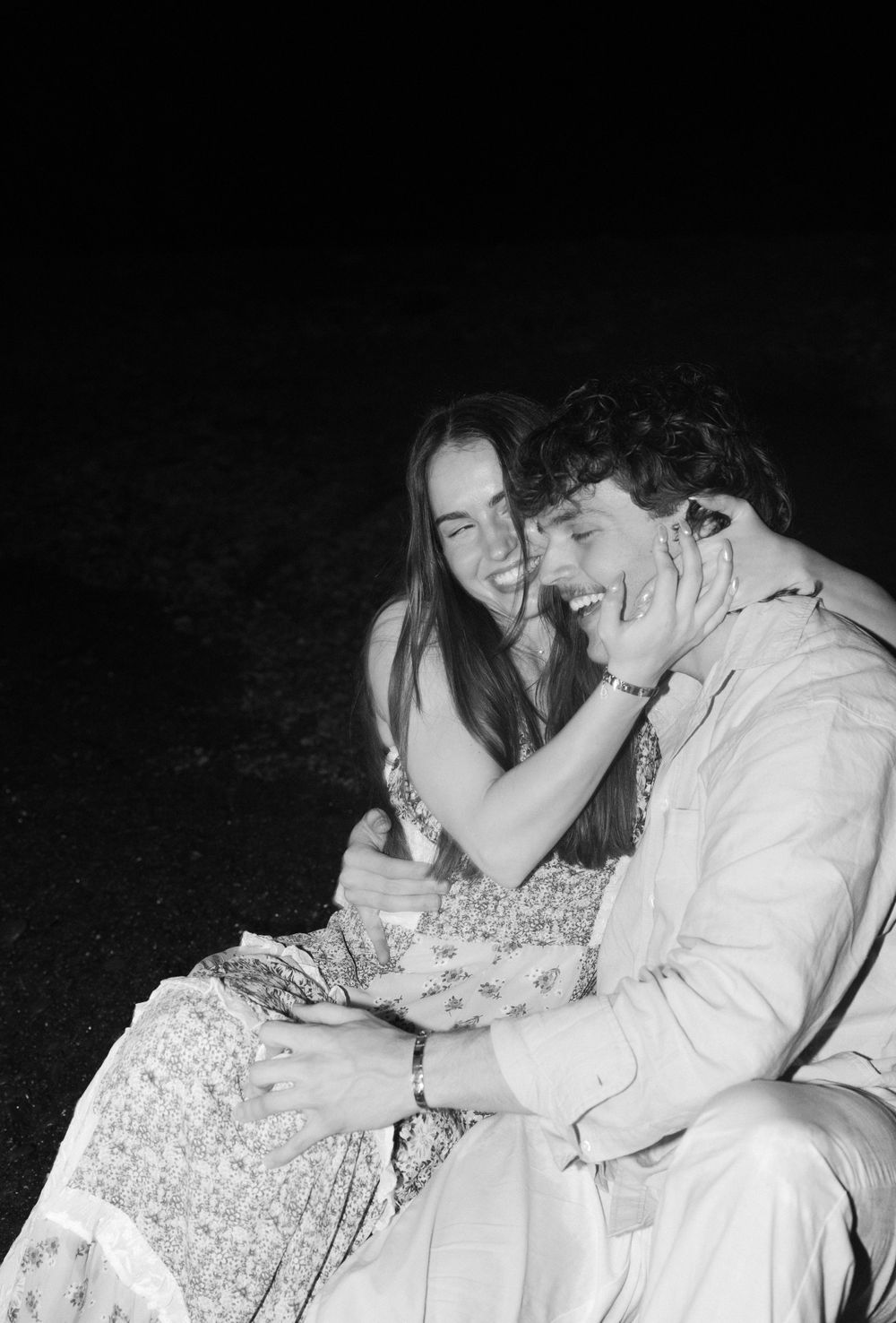 A candid black and white photograph showing a couple laughing together while sitting outside at night.