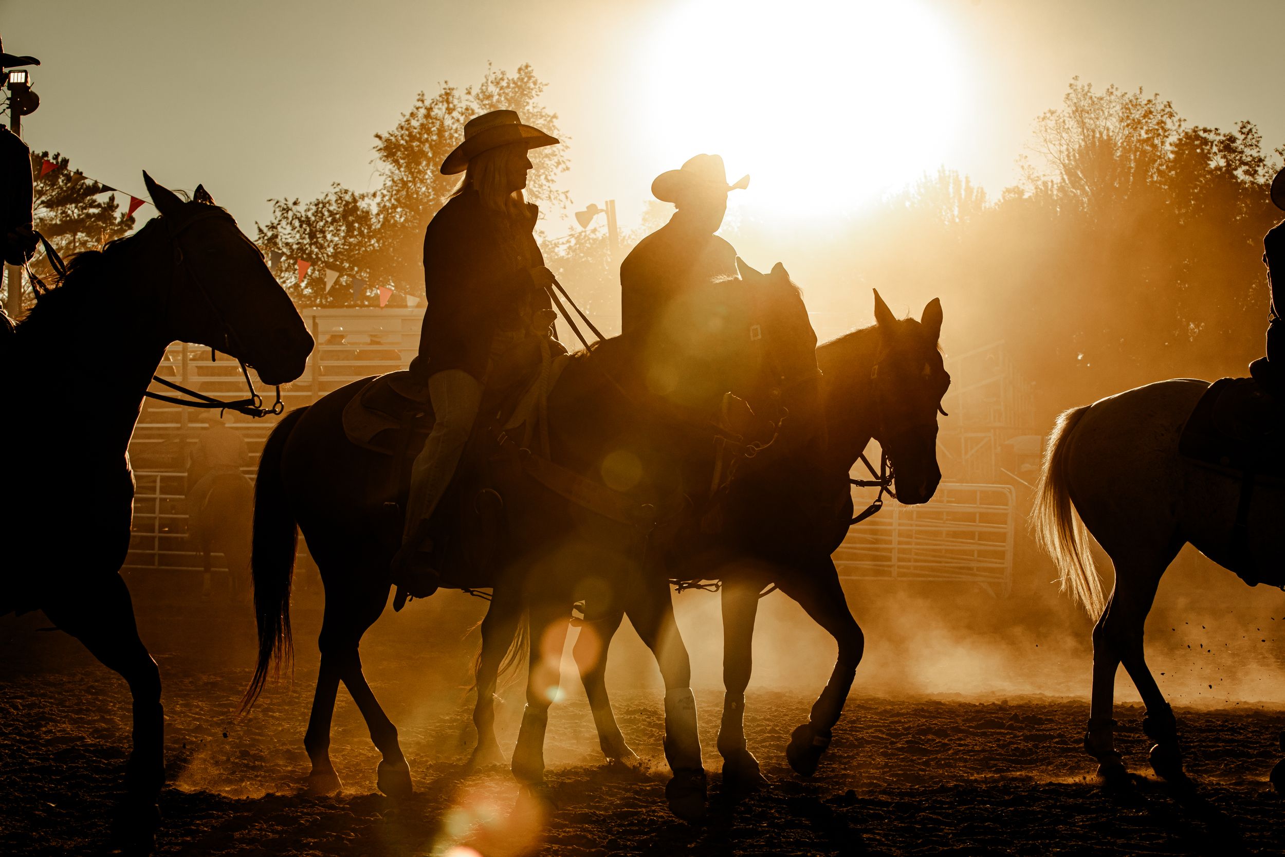 Cowboys riding horses in a dusty arena at sunset casting long shadows.