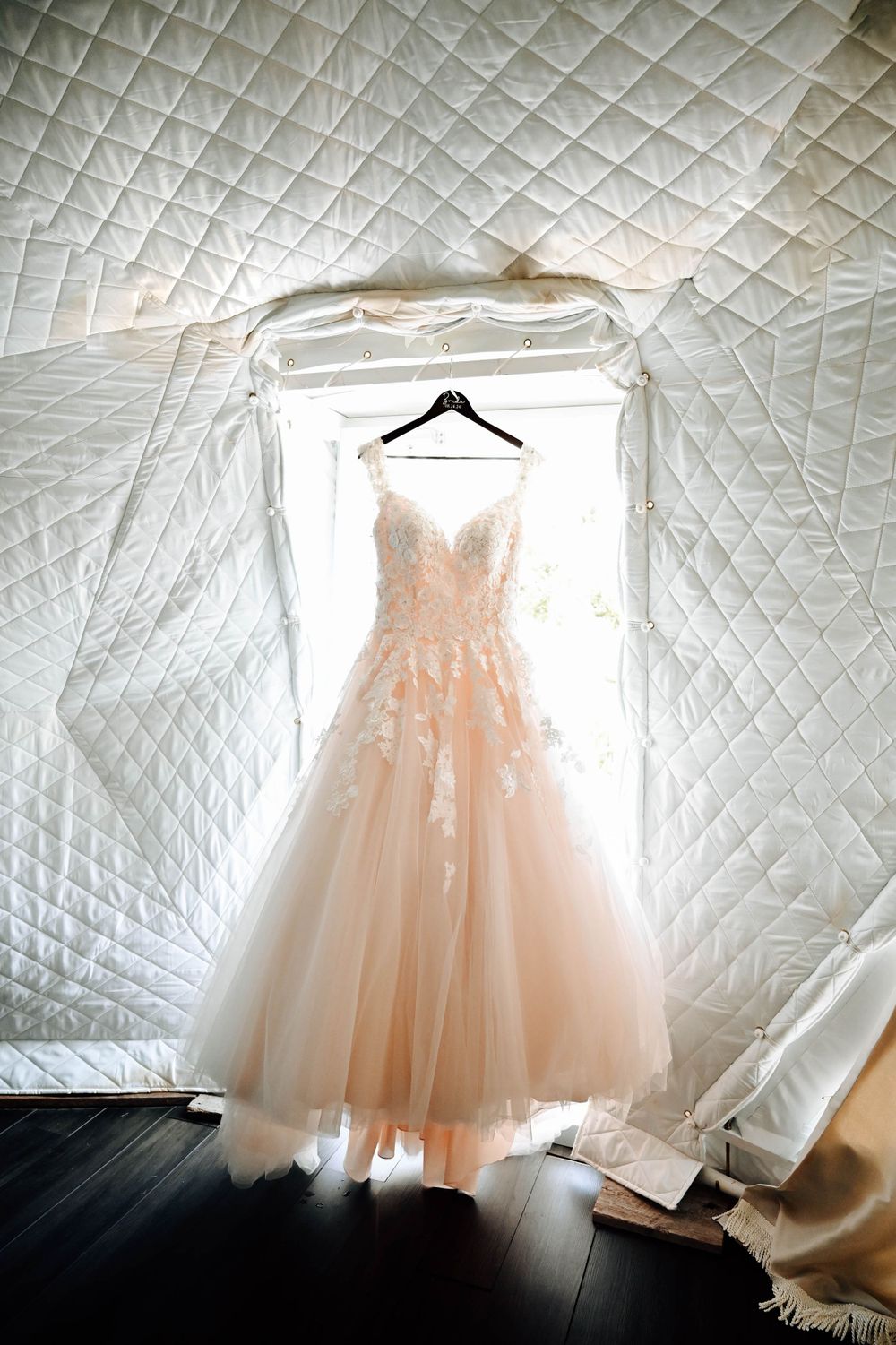 A delicate blush wedding dress with tulle skirt hangs illuminated in a doorway against a quilted white backdrop.