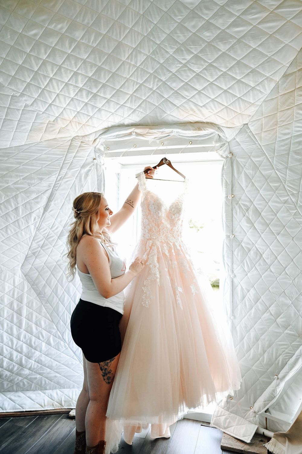 A blush pink tulle wedding gown hangs in front of a white quilted backdrop as someone tends to it.