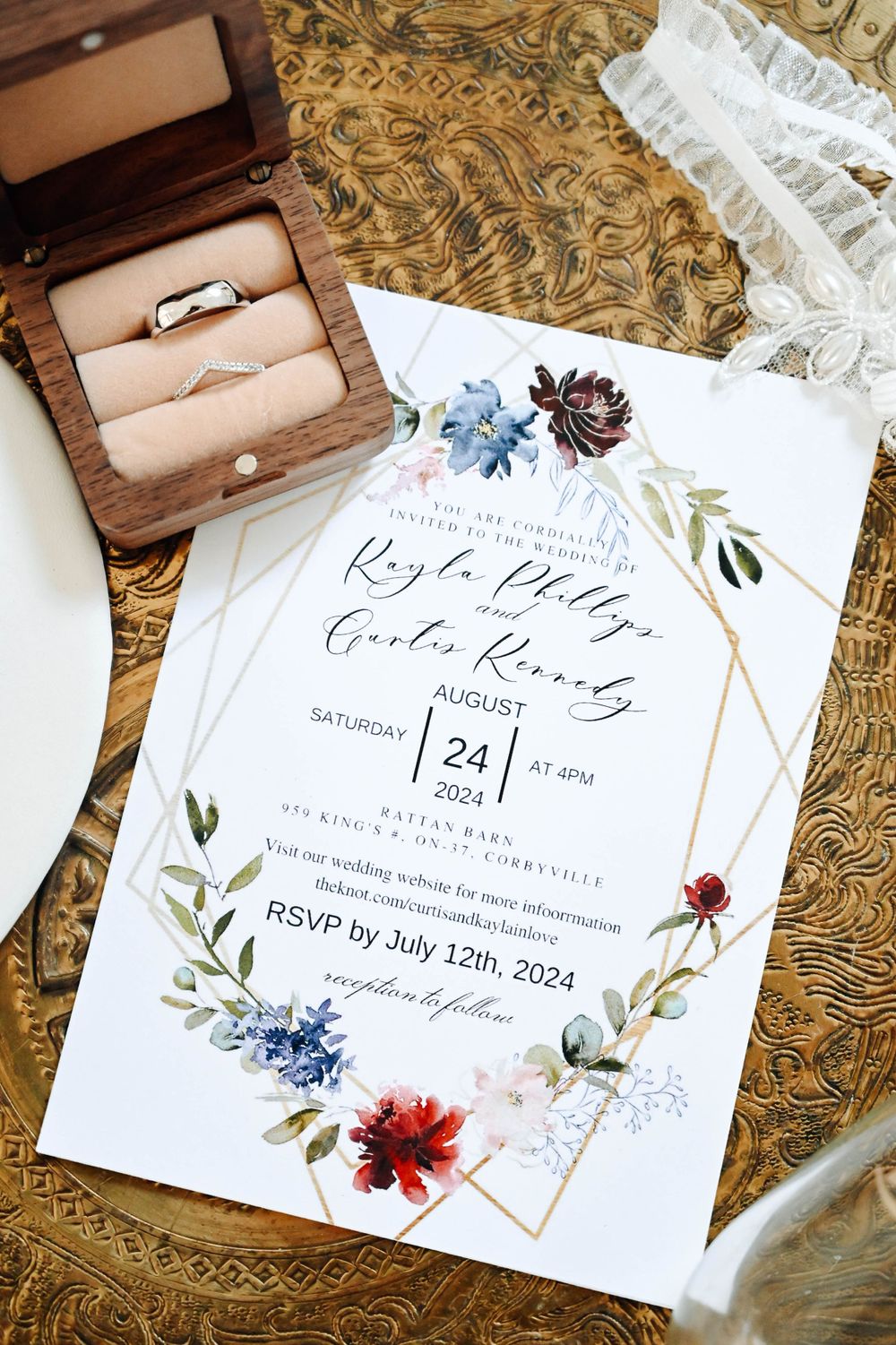 Wedding invitation with floral design and geometric frames next to brown ring box on textured golden fabric background.