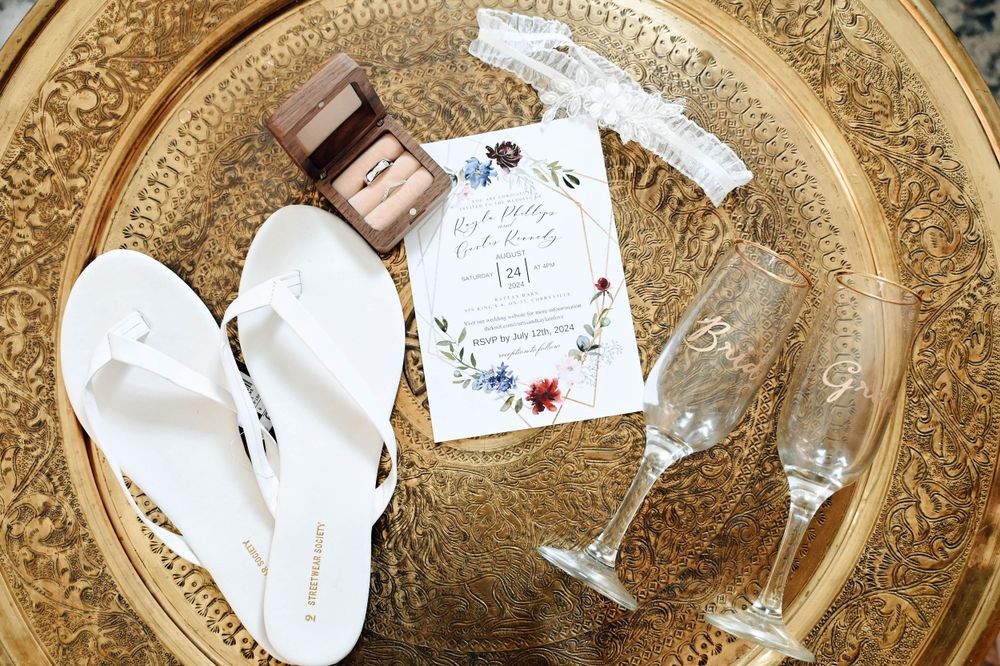 Wedding details including white slippers, champagne glasses and small gift box arranged on ornate gold tray.