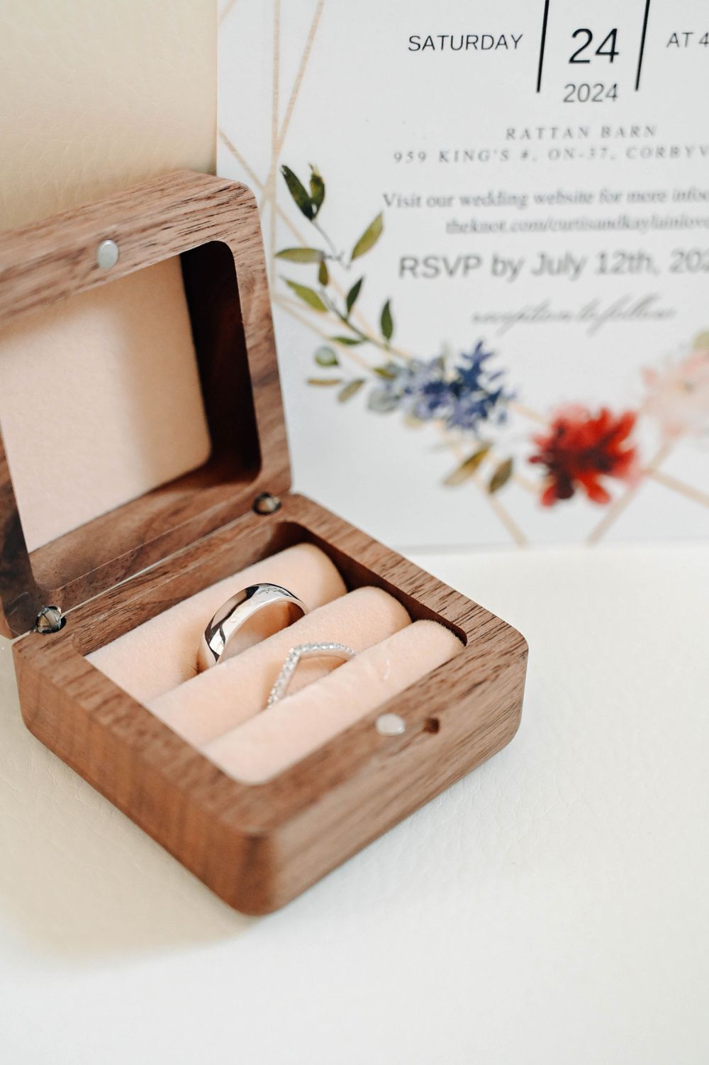 Wedding rings rest in a wooden box next to a floral wedding invitation showing the date Saturday, August 24, 2024.
