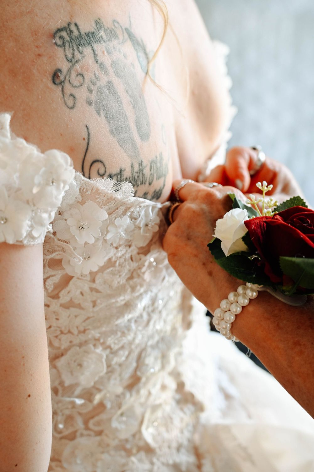 Close-up of a detailed lace wedding dress with visible tattoo while someone pins a red and white floral corsage.