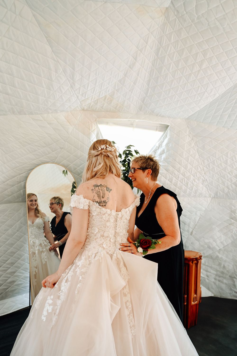 A bride in a lace-back wedding gown gets help with final dress adjustments in front of a mirror.