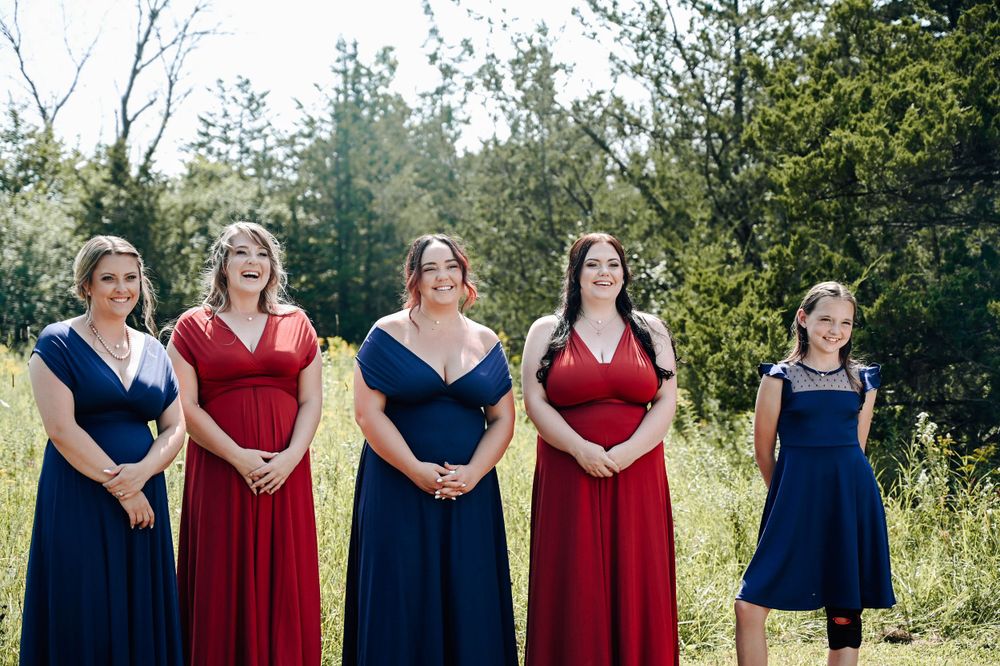 A group of bridesmaids in alternating navy blue and burgundy dresses stand together in a sunny outdoor field.