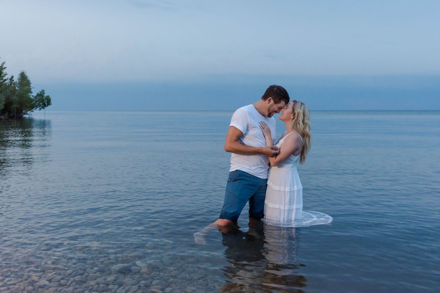 A romantic couple stands in calm lake waters at dusk, embracing while wearing white and blue casual attire.