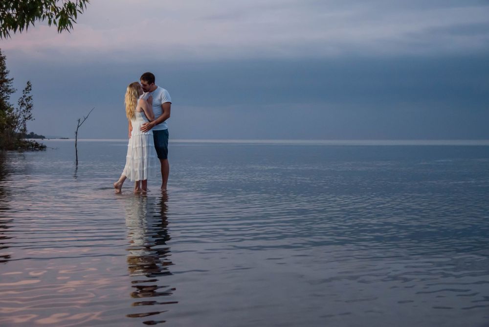 A couple in wedding attire stands together in shallow water at sunset, their reflection rippling in the tranquil water.