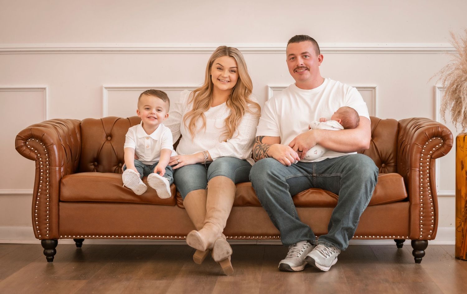 Family sits together on brown leather chesterfield sofa in a light neutral room with pampas grass decor.