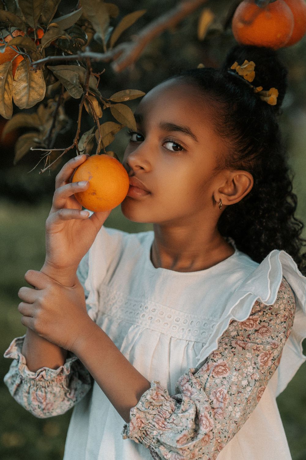 Young child in white lace dress smells fresh orange on tree branch during autumn photo session.