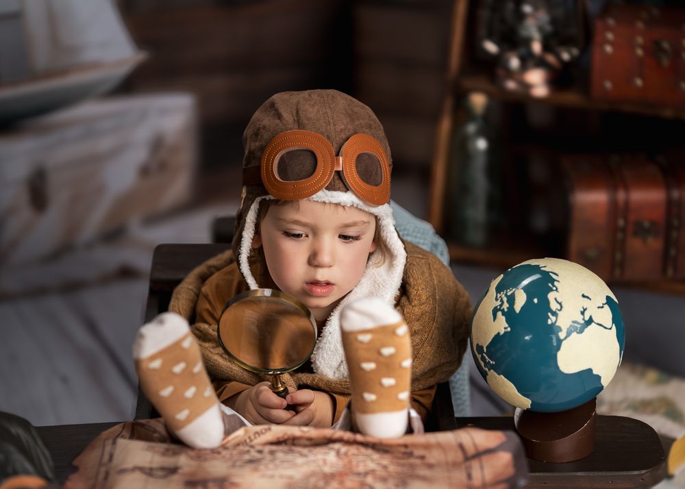 A child in aviator costume looking at vintage globes and maps on a wooden table.
