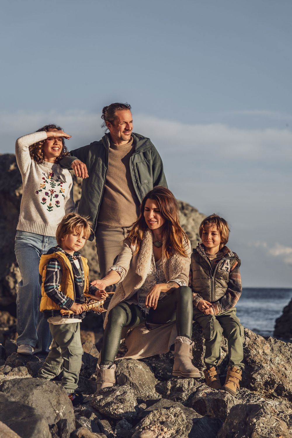 A family in casual clothing sits together on coastal rocks during sunset at the beach.