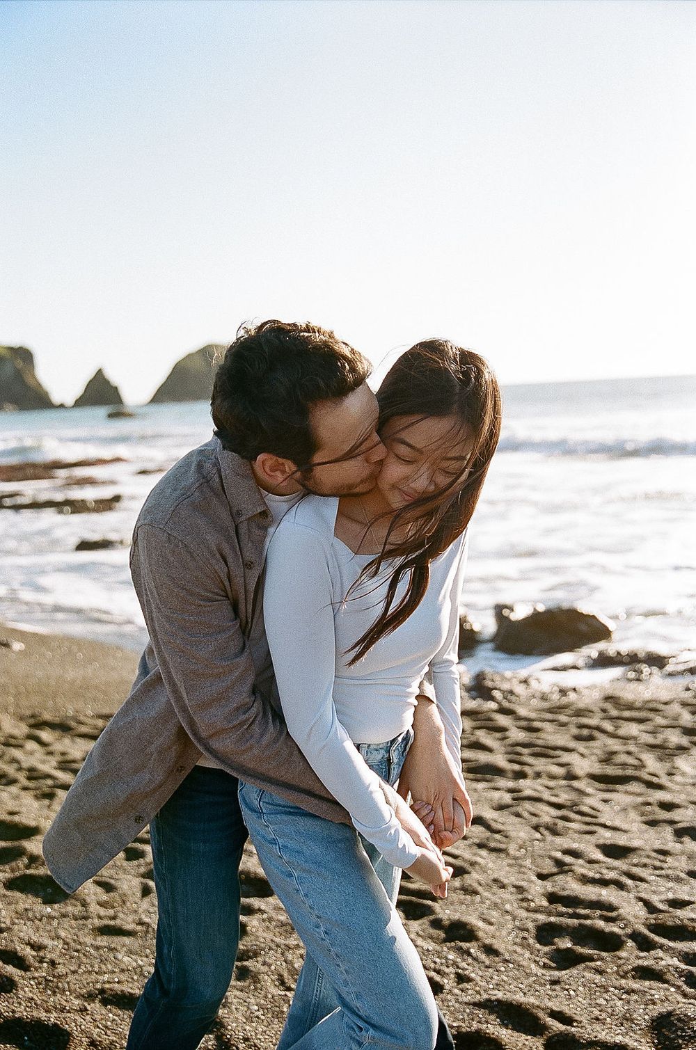 A romantic couple embraces and shares a tender moment together on a sandy beach with rocky coastline in the background.