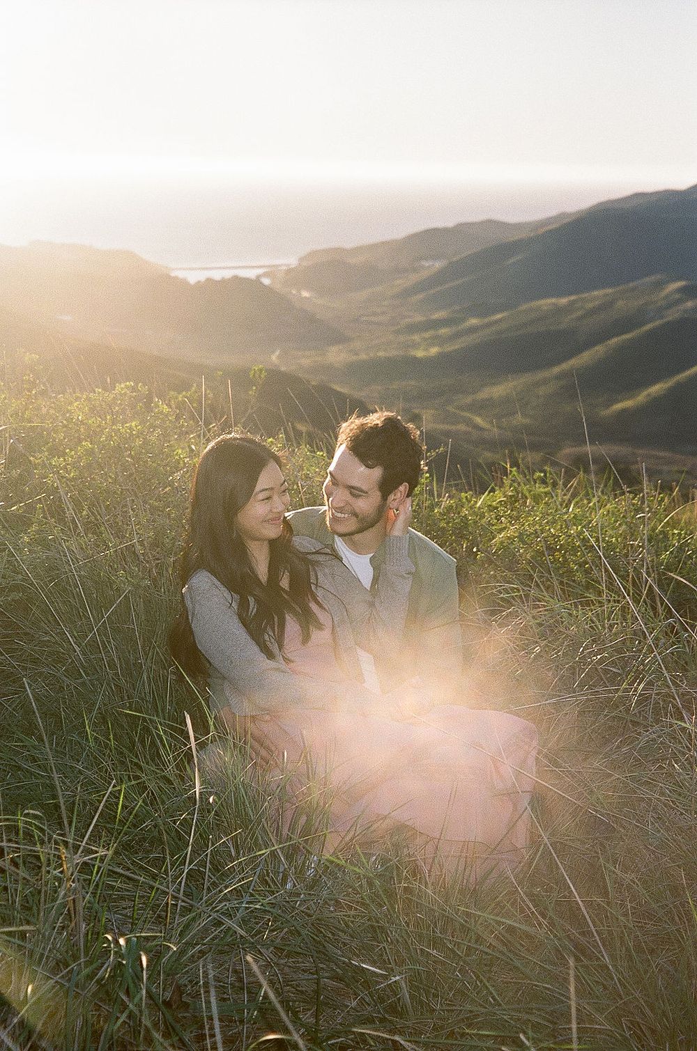 A couple shares an intimate moment at sunset on a grassy hillside overlooking coastal mountains.