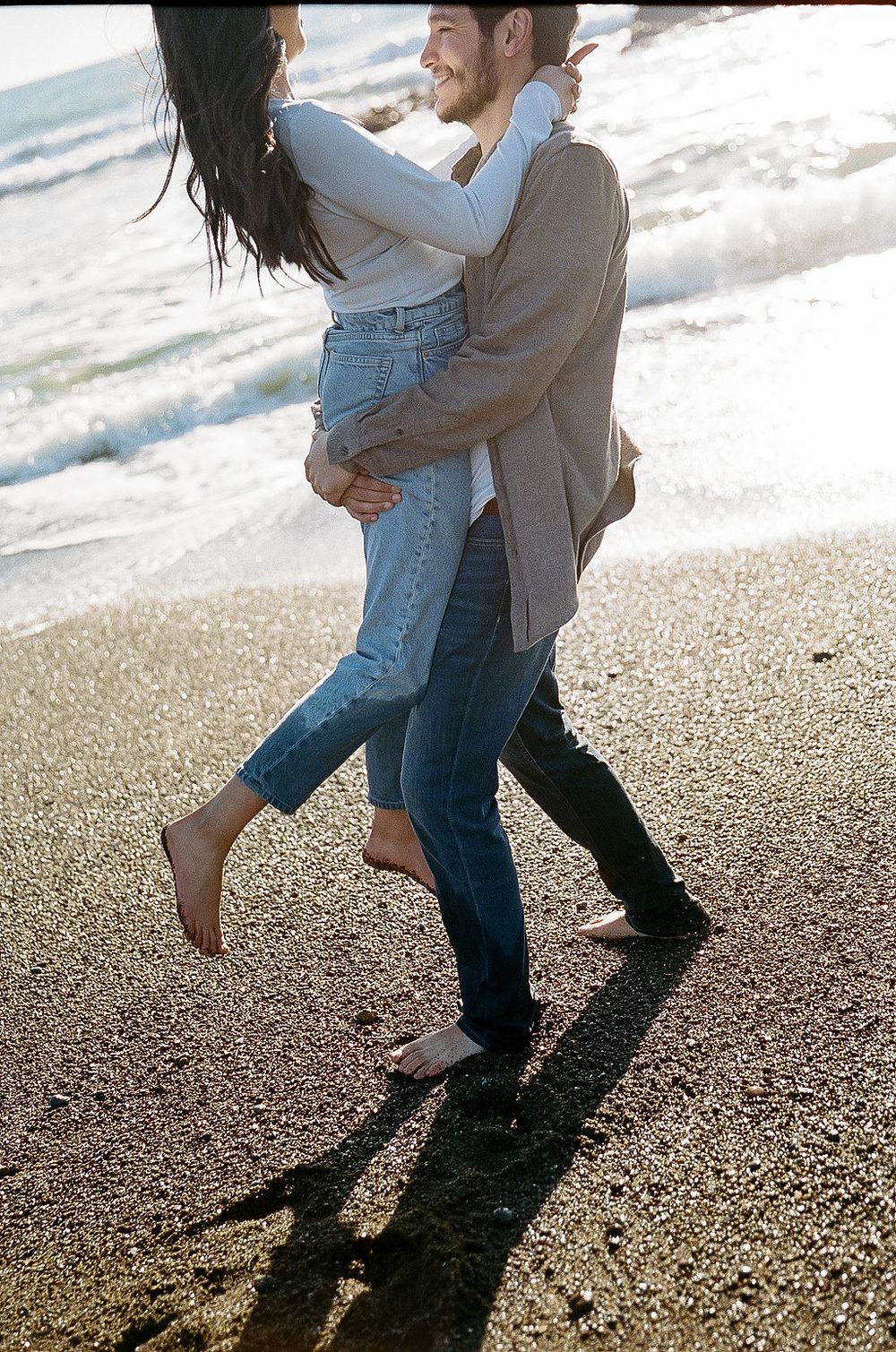 A romantic couple embraces and dances on a sun-lit beach, casting long shadows on the sand.