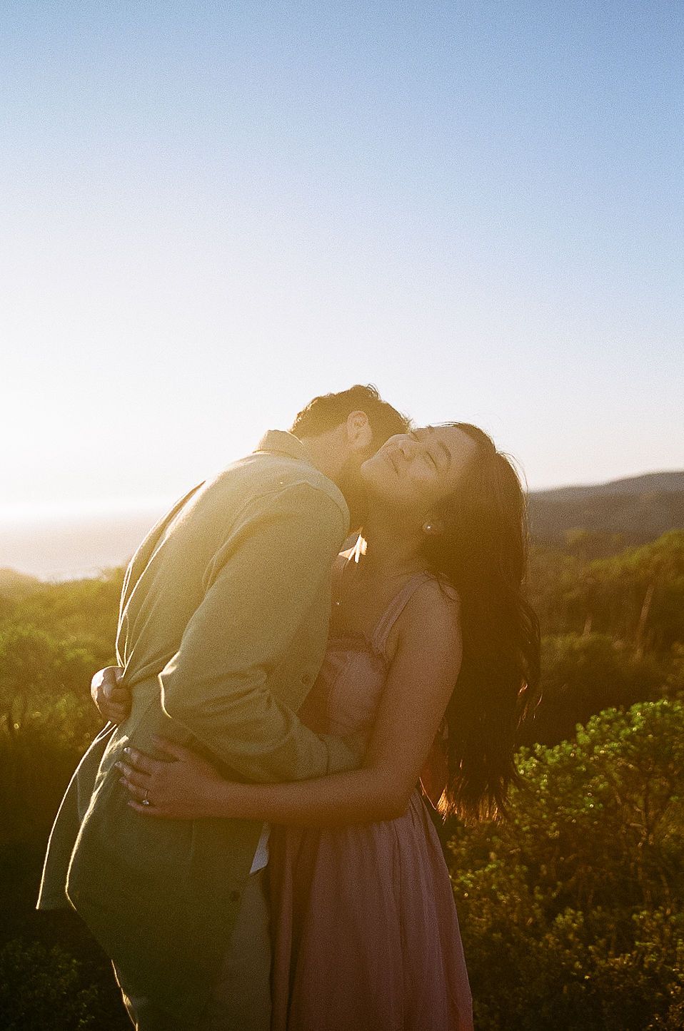 A couple embraces in a warm sunset glow on a hilltop with mountains in the background.