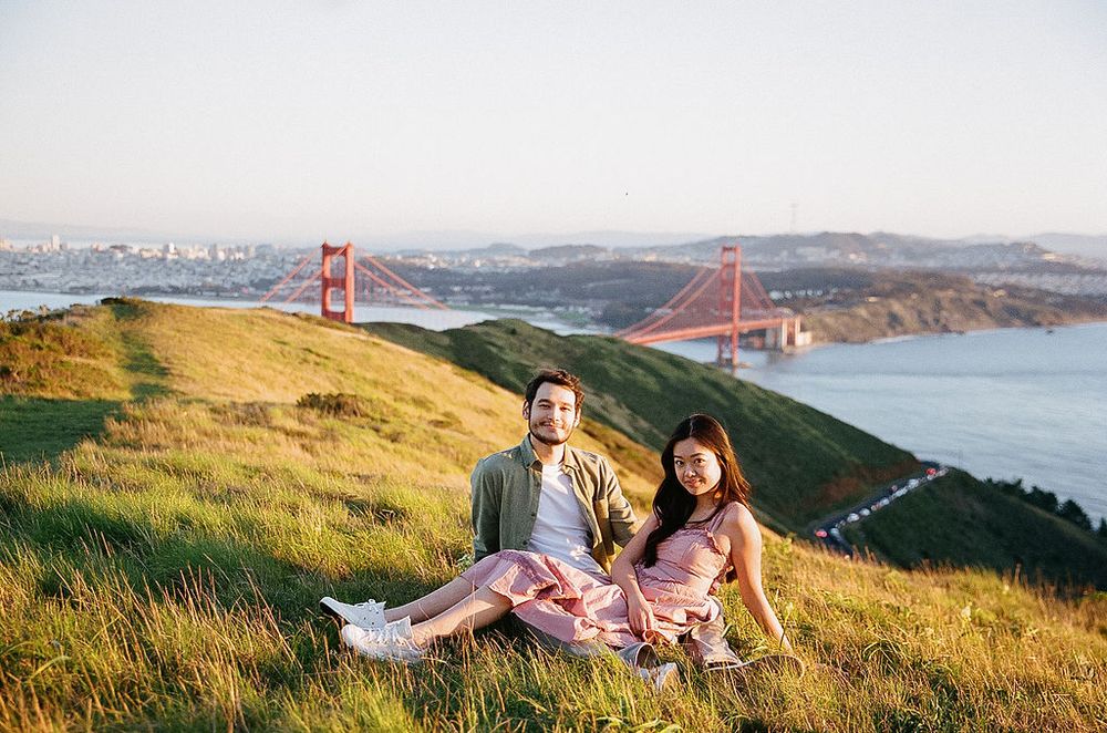 Two people sitting on grassy hill overlooking the Golden Gate Bridge and San Francisco Bay on a sunny afternoon.