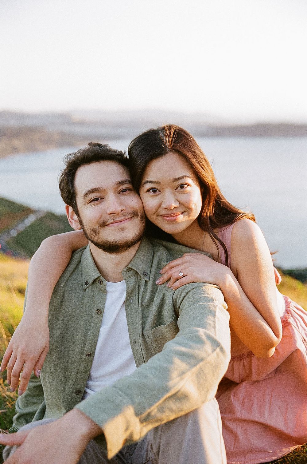 A happy couple embraces while sitting outdoors during golden hour with scenic landscape in the background.