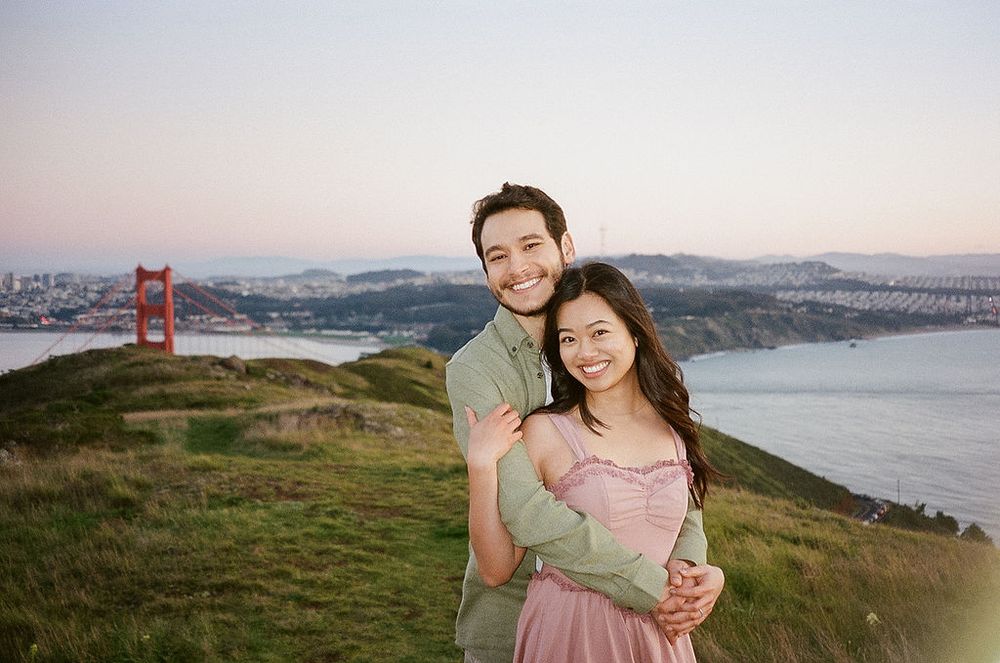 A couple poses on a hilltop overlooking the Golden Gate Bridge and San Francisco Bay during sunset.