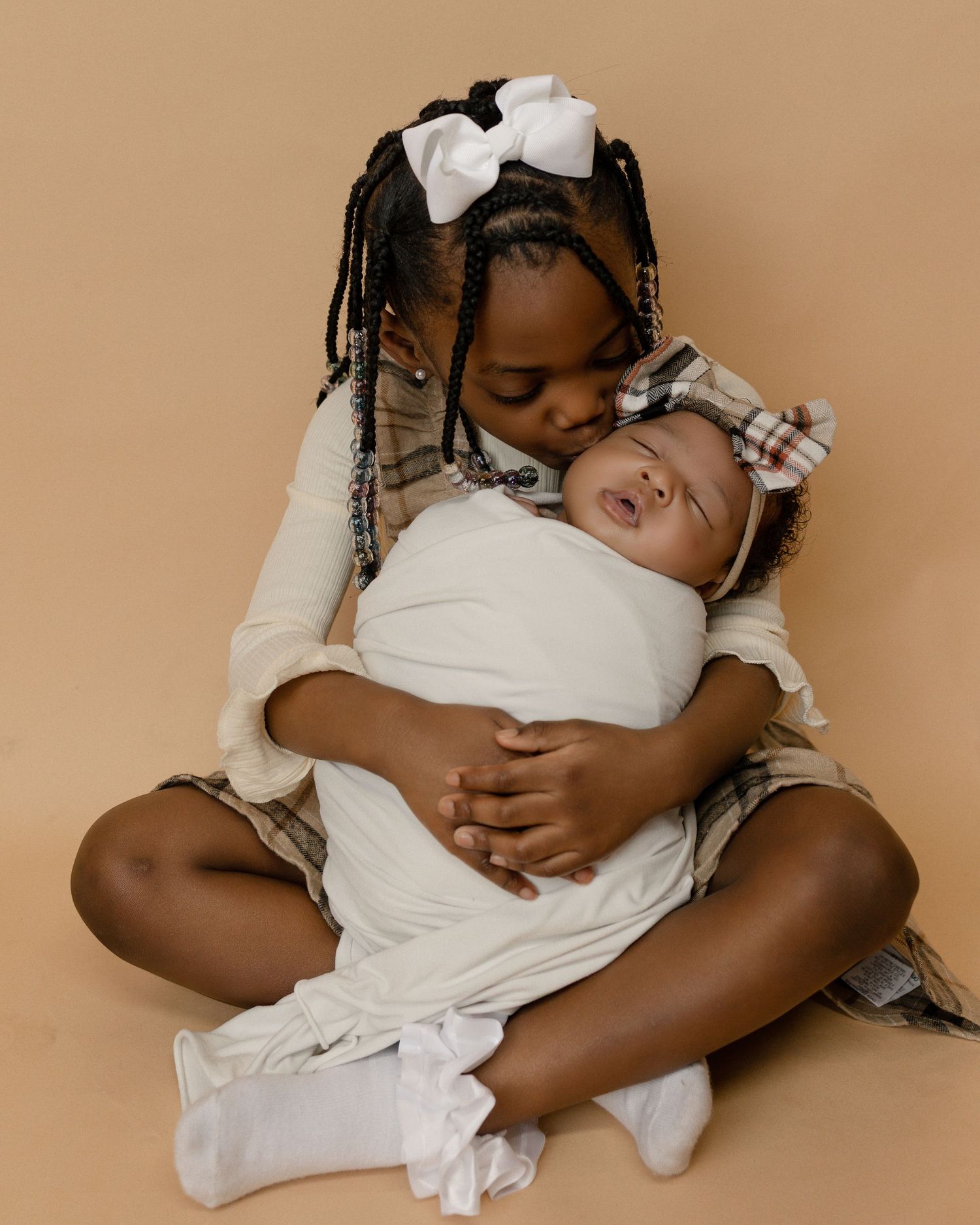Two children in white outfits with bows in their hair embrace tenderly on a beige backdrop.