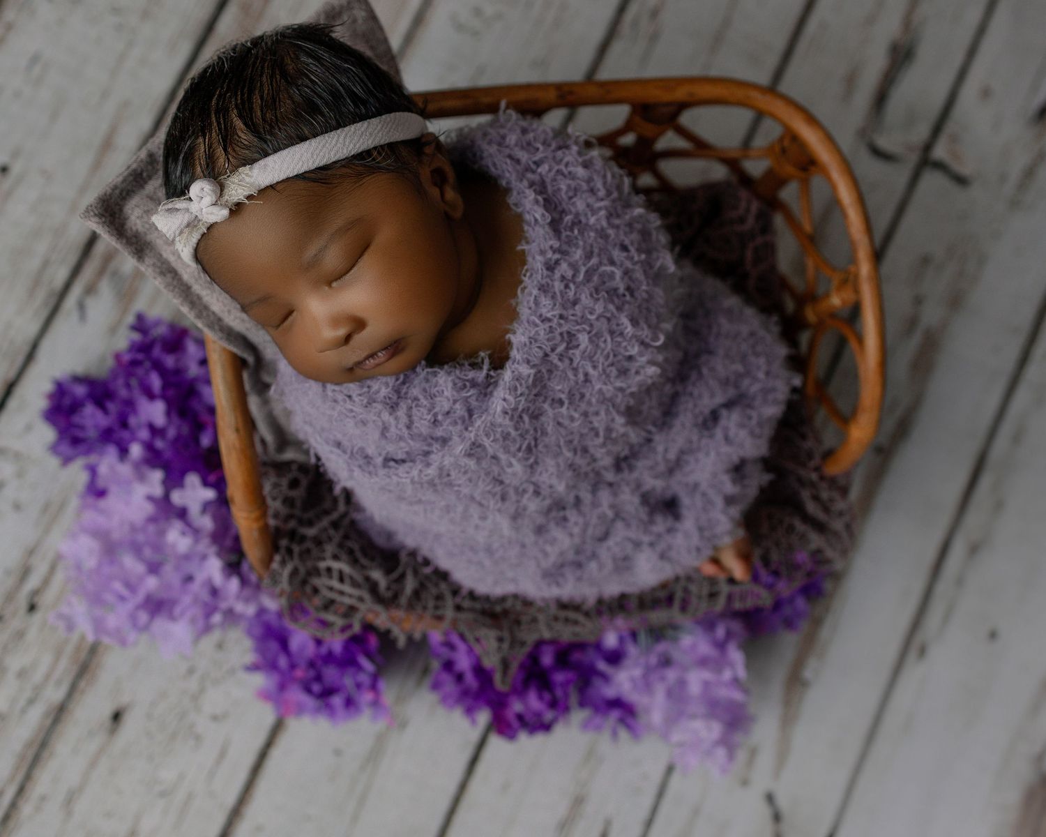 Newborn baby sleeping peacefully in wooden basket with purple flowers on white wooden floor.