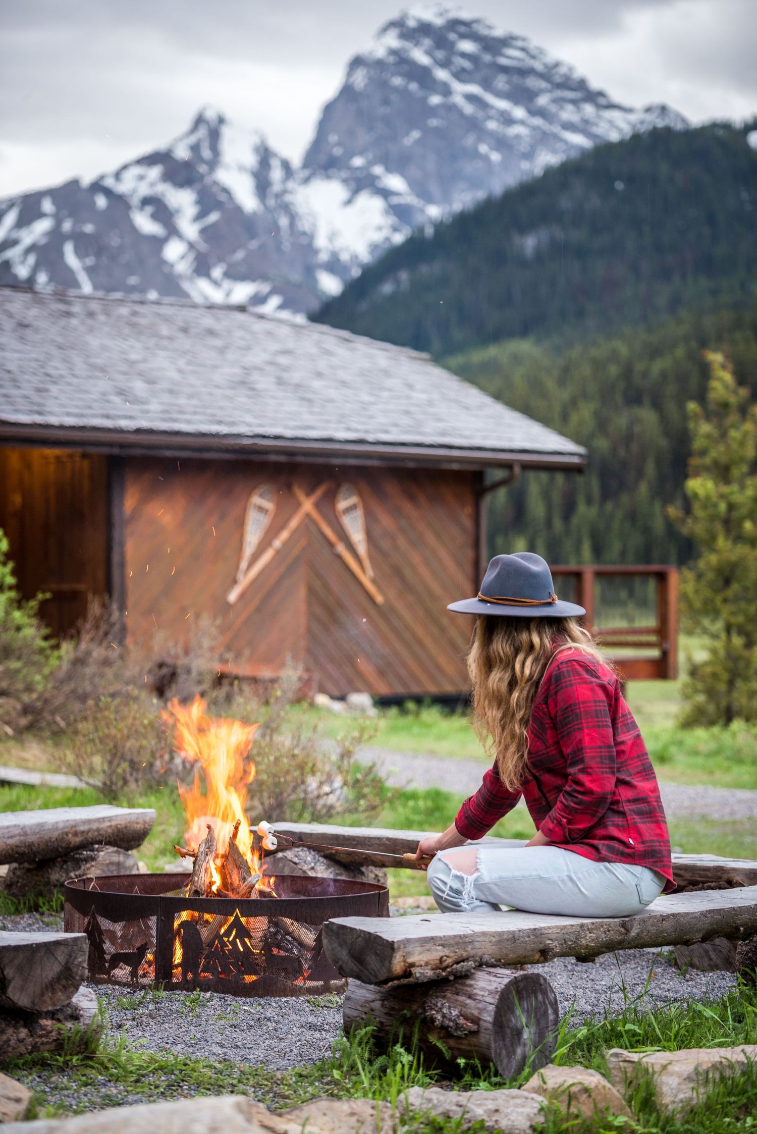 Person in plaid shirt and hat relaxes by campfire at rustic cabin with snow-capped mountains in background.