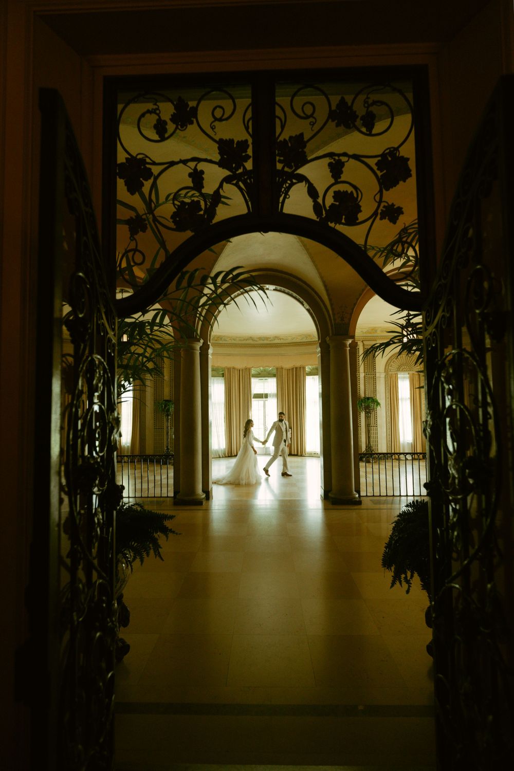 Silhouetted figures in formal attire viewed through an ornate archway with decorative ironwork and greenery.