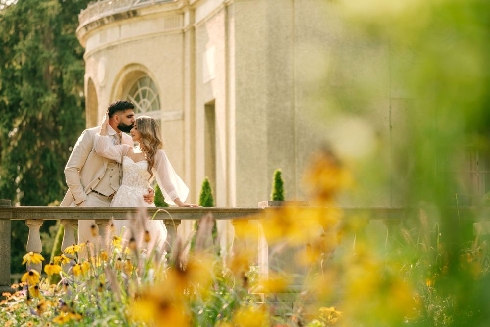 Couple shares romantic moment on stone balcony with yellow wildflowers in foreground and classical architecture behind them.